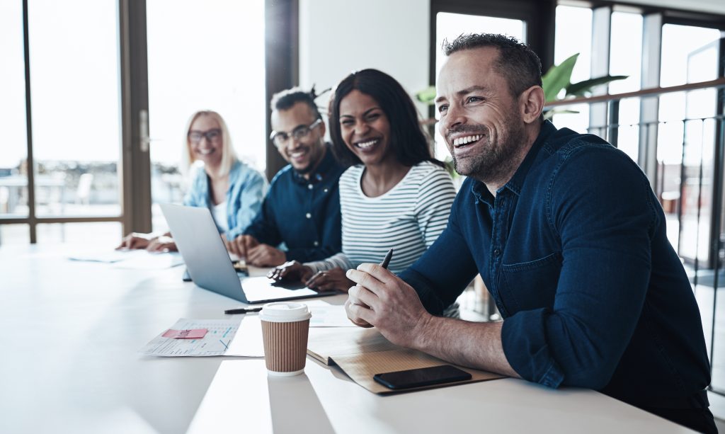 Diverse group of young businesspeople laughing while sitting together in a row at an office desk during a meeting